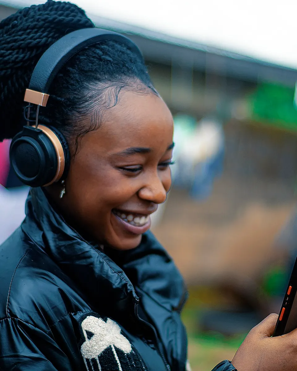 A young woman with headphones smiling at her phone outdoors